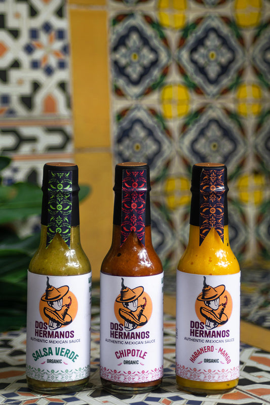Three bottles of hot sauce with decorative labels, each filled with a different colored sauce, displayed on a surface with a patterned background.