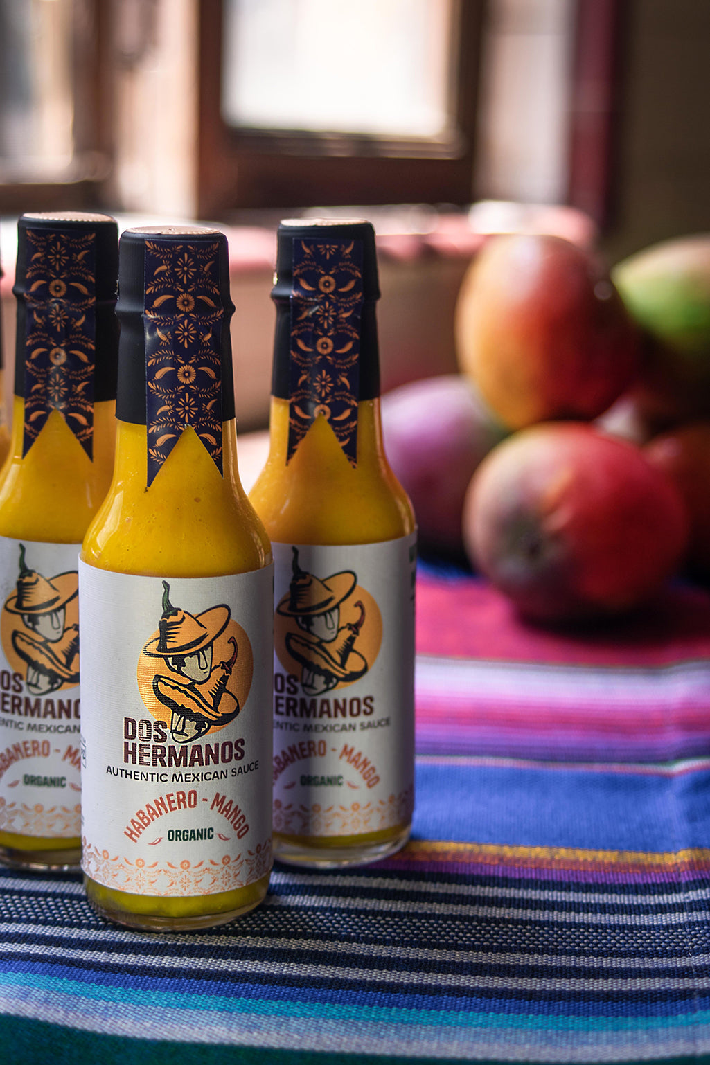 Three bottles of Dos Hermanos organic hot sauce with habanero and mango flavor, placed on a surface with a colorful striped cloth and fruits in the background.