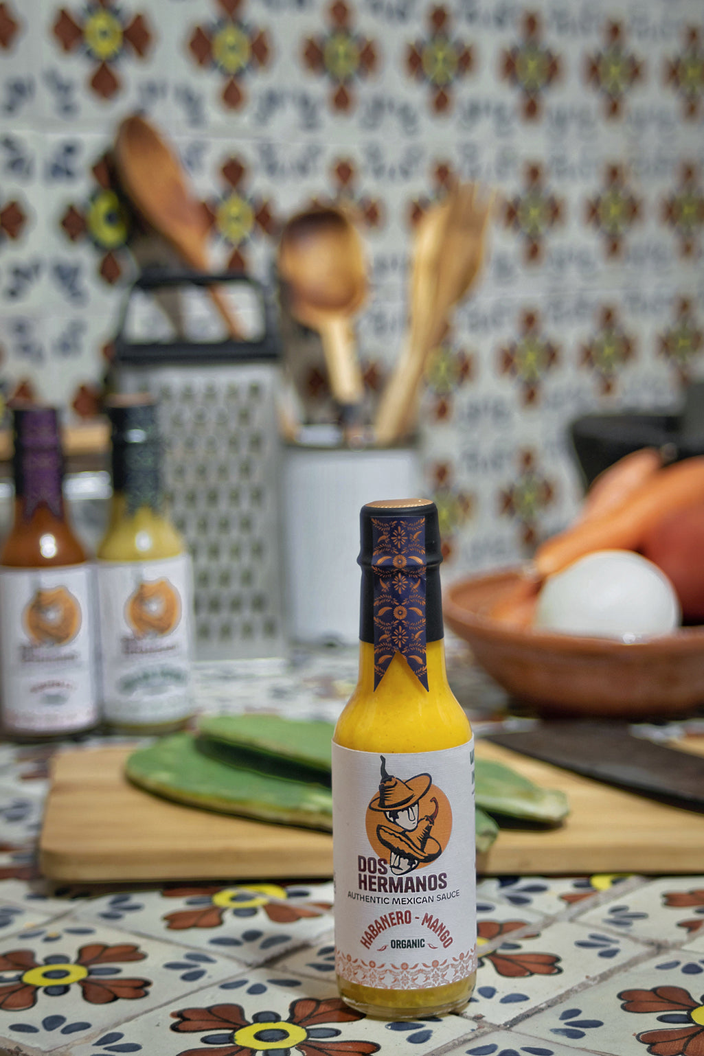 A bottle of Organic Habanero-Mango hot sauce displayed on a table with a colorful patterned background, surrounded by various kitchen items.