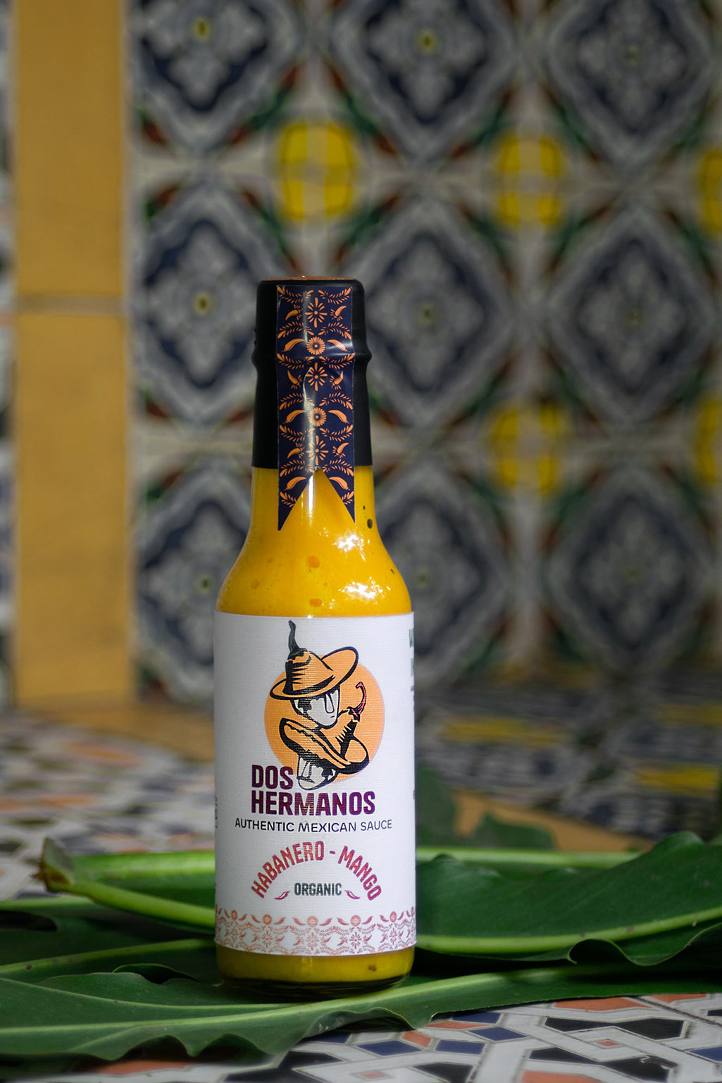A bottle of Organic Habanero-Mango hot sauce displayed on a table with a colorful patterned background, surrounded by various kitchen items.