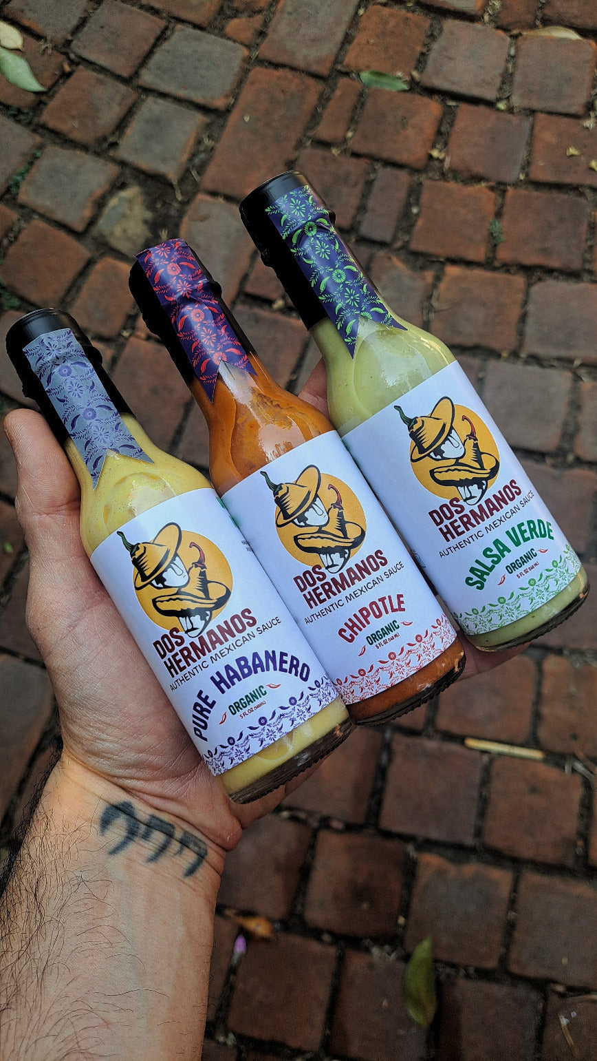 Hand holding three bottles of Dos Hermanos hot sauce on a brick surface