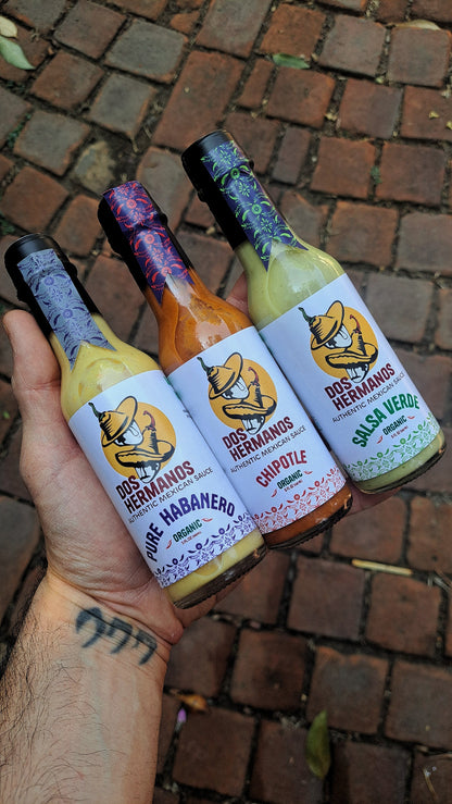 Hand holding three bottles of Dos Hermanos hot sauce on a brick surface