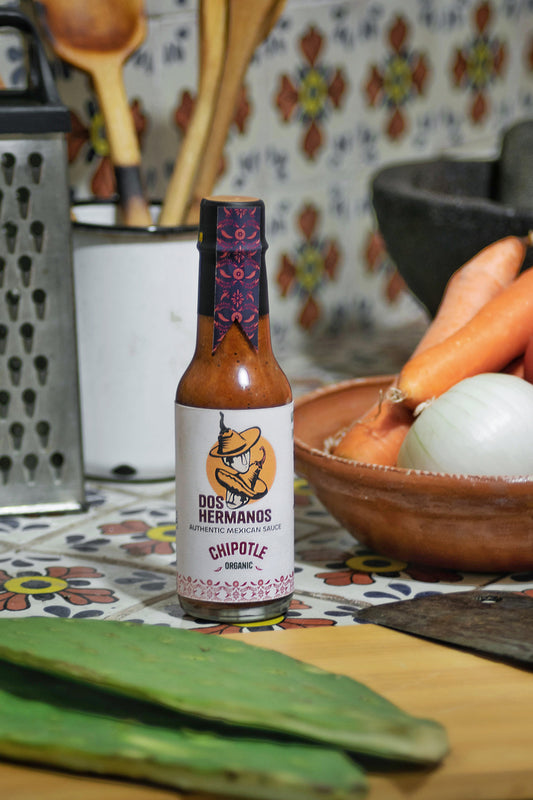 A bottle of Organic Chipotle sauce with a label featuring a rooster, placed on a kitchen counter surrounded by vegetables.