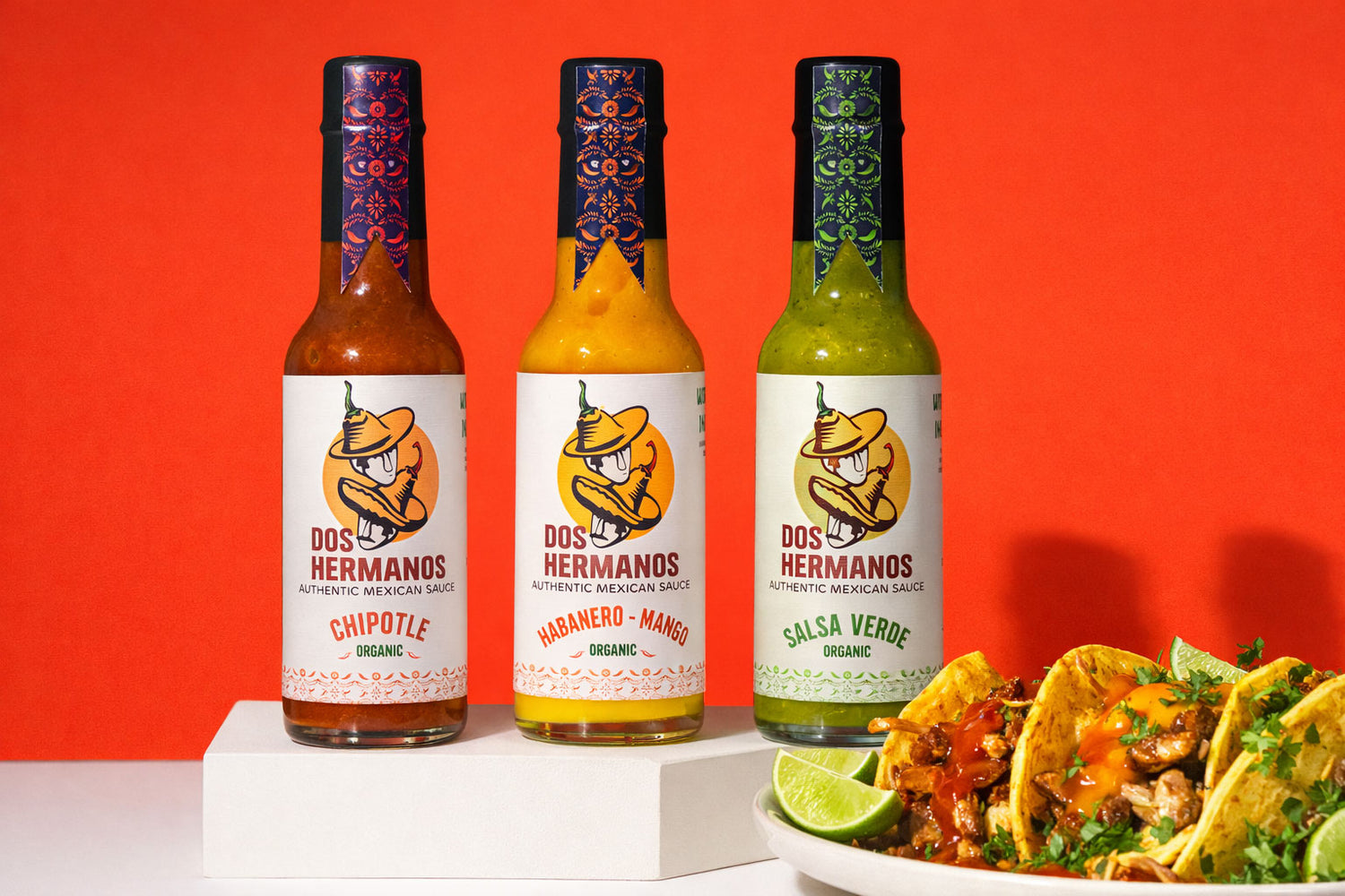 Three bottles of Dos Hermanos hot sauce with a plate of tacos on a red background