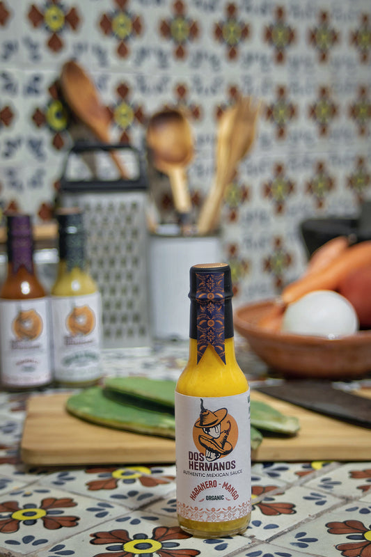 A bottle of Organic Habanero-Mango hot sauce displayed on a table with a colorful patterned background, surrounded by various kitchen items.