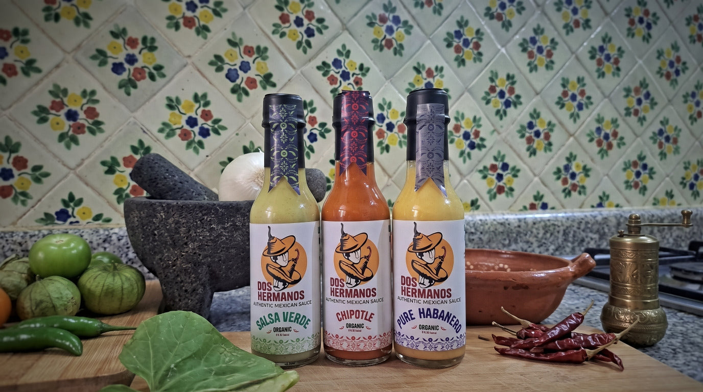 Three bottles of Dos Hermanos hot sauce lined up on a wooden cutting board in a kitchen setting, surrounded by fresh ingredients like tomatoes, chilies, and herbs. The background features a decorative tile wall.