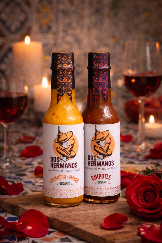 Two bottles of Dos Hermanos hot sauce on a wooden board with candles and wine glasses in the background.