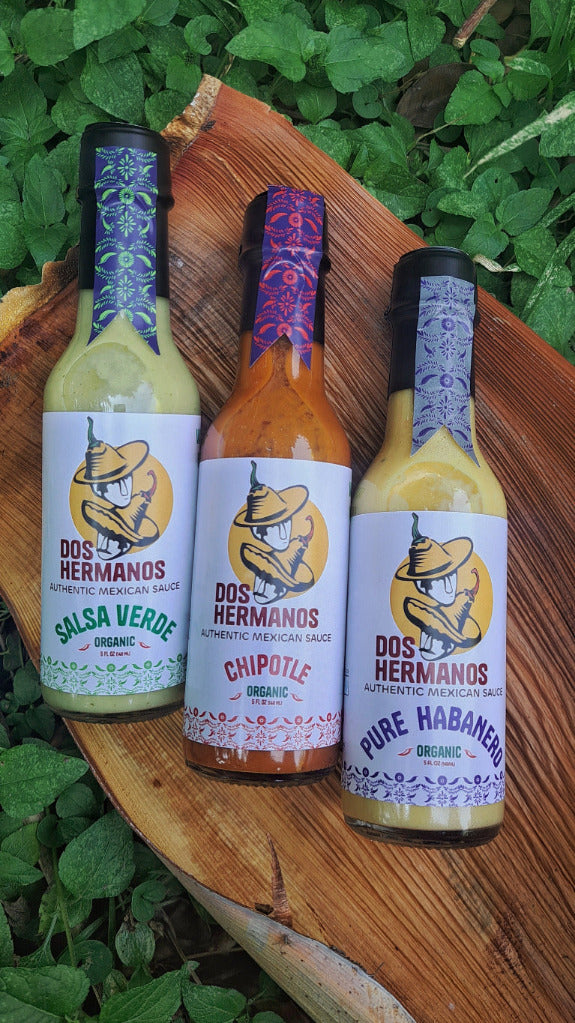 Three bottles of Dos Hermanos hot sauce on a wooden board with greenery in the background