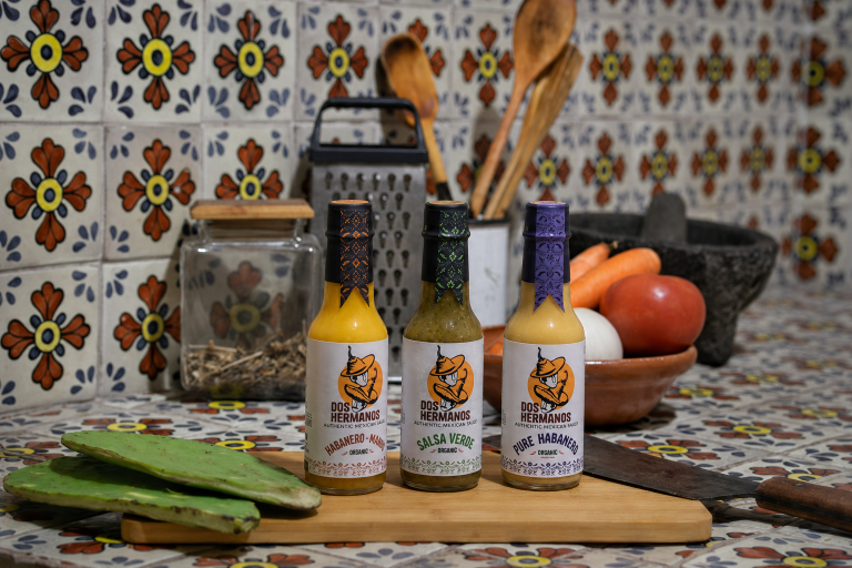 Three bottles of 'Los Hermanos' hot sauce on a wooden cutting board with kitchen items in the background.