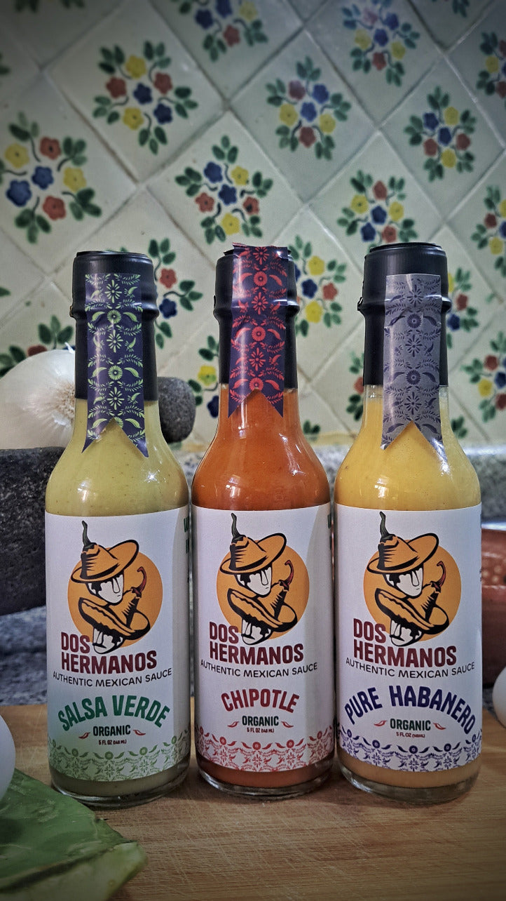 Three bottles of Dos Hermanos salsa on a wooden surface with a colorful tiled wall background.