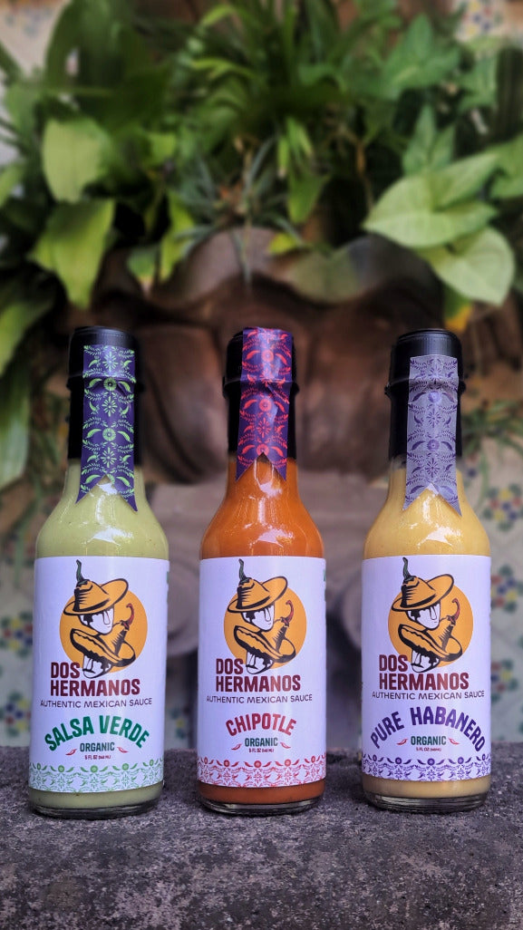 Three bottles of Dos Hermanos hot sauce with labels on a dark surface and green plants in the background.