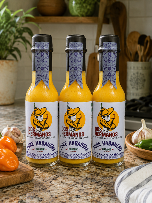 Three bottles of Dos Hermanos Pure Habanero sauce on a kitchen counter with ingredients in the background.
